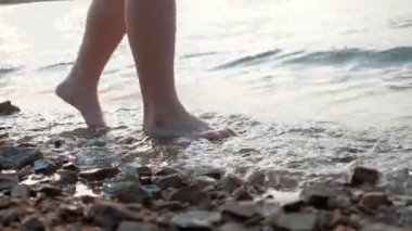 Arka planda okyanus dalgaları olan kumsalda çakıl taşı kumlarında yürüyen kadın ayaklarına yakın çekim. Çakıl taşlı kuma basan kadın bacakları. Deniz kıyısındaki çıplak ayaklı kız. Yaz tatili.