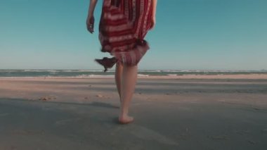 Kumsalda çıplak ayakla yürüyen ağır çekimli kadın rüzgarda esen uzun elbiseyle kumsalda ayak izleri bırakıyor. Yaz zamanı tatil..