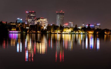BUCHAREST, ROMANIA - 30 Ekim 2021 Herastrau Parkı 'ndan gölün suyundaki şehir ışıklarının renkli yansımasının gece görüntüsü. 