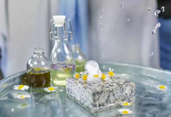 Handmade cosmetics. Shooting on the table at an angle of 90 degrees. Bottles of butter and perfume on a table of water. On a granite stone can of cream, around fresh chamomile flowers