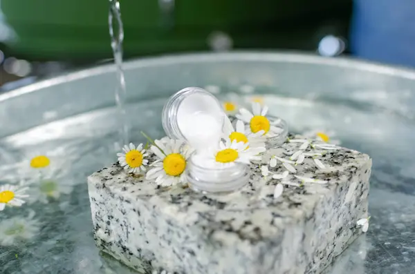 Handmade cosmetics. Shooting on the table at an angle of 90 degrees. Bottles of butter and perfume on a table of water. On a granite stone can of cream, around fresh chamomile flowers