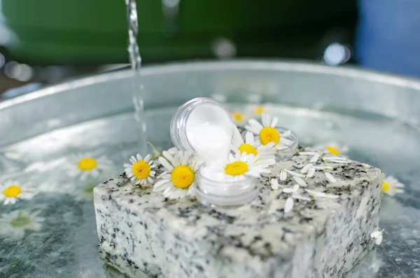 Handmade cosmetics. Shooting on the table at an angle of 90 degrees. Bottles of butter and perfume on a table of water. On a granite stone can of cream, around fresh chamomile flowers