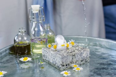 El yapımı kozmetik. Masaya 90 derecelik bir açıyla ateş ediyorlar. Su masasında tereyağı ve parfüm şişeleri. Taze papatya çiçeklerinin etrafındaki granit bir krema kutusunda.