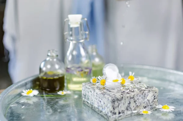 Handmade cosmetics. Shooting on the table at an angle of 90 degrees. Bottles of butter and perfume on a table of water. On a granite stone can of cream, around fresh chamomile flowers