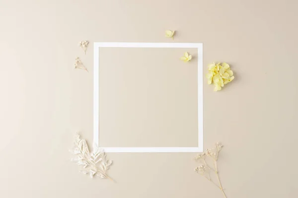 Cosmetic background with flowers and white frame on pastel beige. Flat lay, copy space