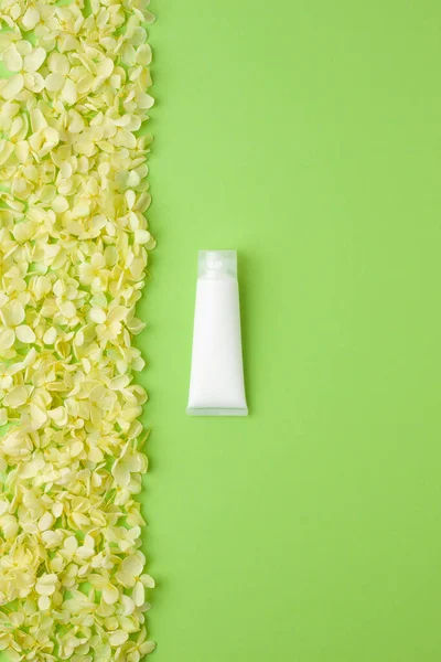 White tube of cosmetic cream with flowers on green background. Flat lay, copy space