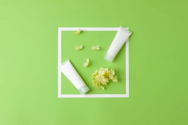 White tubes of cosmetic cream with white frame and flowers on green background. Flat lay, copy space