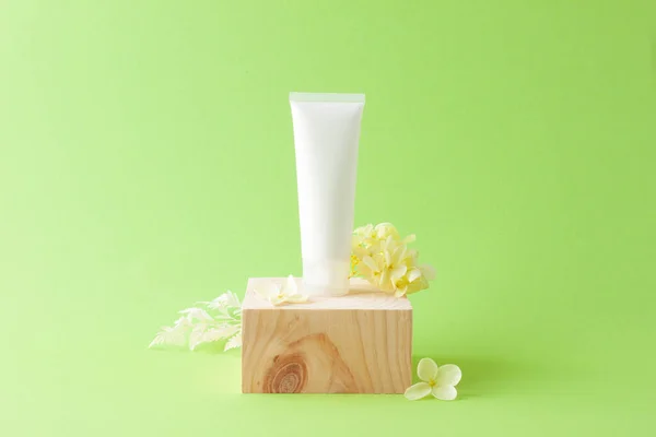 White tube of cosmetic cream with wood podium and flowers on green background. Close up, copy space