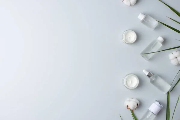 Cosmetic skin care products with flowers and palm leaf on grey background. Flat lay, copy space