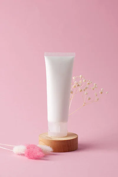 White tube of cosmetic cream with flowers on wooden podium on pink background. Close up.