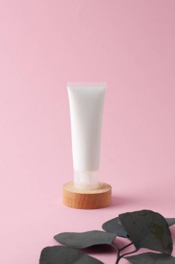 Beyaz kozmetik krem tüpü, okaliptüs ve pembe arka planda tahta podyum. Kapat..