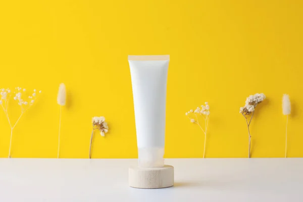 White tube of cosmetic cream with flowers on wooden podium on yellow background. Close up.