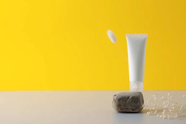 White tube of cosmetic cream on a stone podium with flowers on yellow background. Close up, copy space.