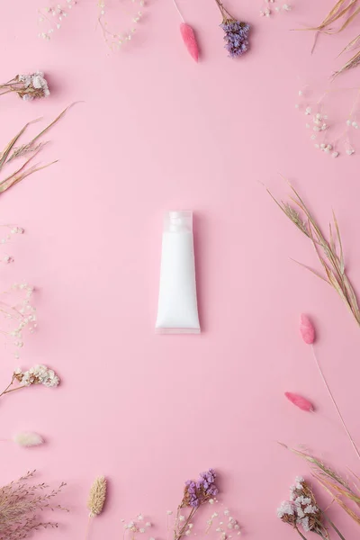 White tube of cosmetic cream with flowers on pink background. Flat lay