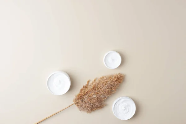 Jars of cosmetic cream with pampas on pastel beige background. Flat lay, copy space