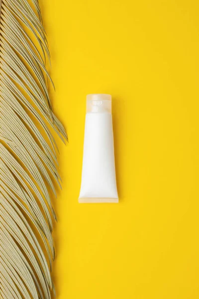 White tube of cosmetic cream with palm leaf on yellow background. Flat lay
