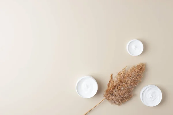 Jars of cosmetic cream with pampas on pastel beige background. Flat lay, copy space