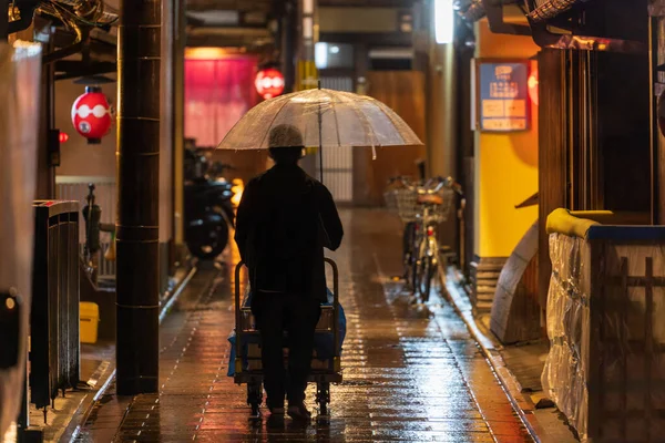 Człowiek z parasolem popycha wózek przez deszczową uliczkę w historycznej dzielnicy w nocy — Zdjęcie stockowe
