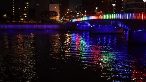 Colorful bridge reflects off ripples in river through city at night — Vídeos de Stock