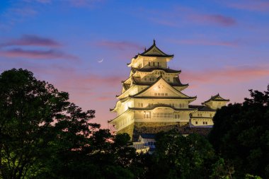 Blue evening sky mixes with sunset color as moon sets over Himeji Castle. High quality photo