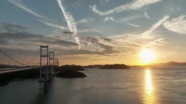 Long suspension bridge with many towers through small island as sun sets into sea. High quality 4k footage