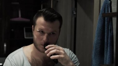 Bir adam bıyığını ve sakalını budayarak, sinematikle, banyodaki aynanın önünde genç bir adamla tıraş ediyor.