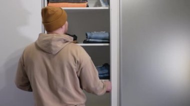 Genç bir adam dolaptan kot pantolon çıkarır, günlük kıyafet seçer. Giysilerinizi dolaptan seçin..