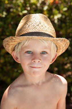 Mavi gözlerle gülümseyen şişkin saçlı çocuğun portresi ve bulanık yaz yeşili arka planlı hasır şapka.