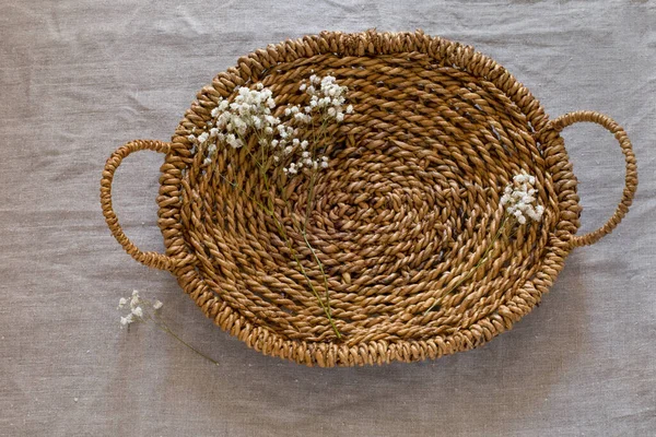Top view of brown woven basket with two handles and tiny white flowers on linen fabric background