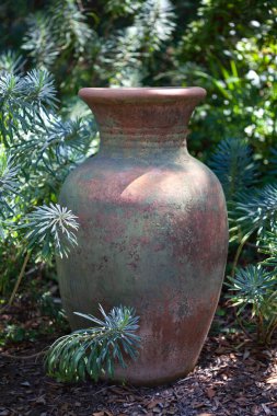 Large decorative clay textured vase on the ground in the garden with blurred greens background