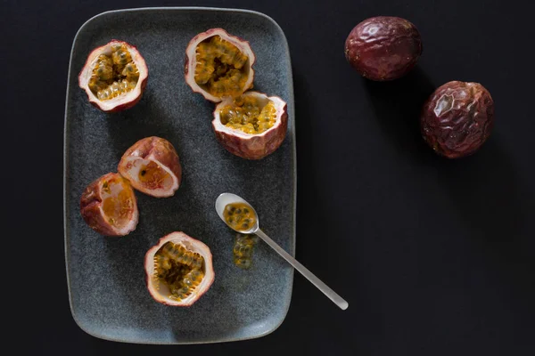 Çarkıfelek meyvesinin en üst görüntüsü ve ikiye bölünmüş, mavi seramik tabakta, metal bir çay kaşığıyla, koyu arkaplanda biraz hamuru dökülmüş.