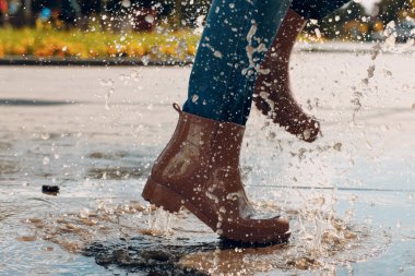 Kauçuk çizme giyen bir kadın sonbahar yağmurunda su birikintisine atlayıp koşuyor.