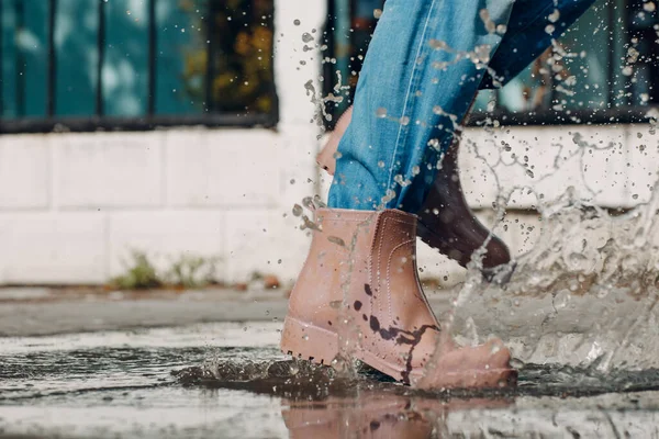 Жінка в дощових гумових черевиках ходить і стрибає в калюжу з водяними бризками і падає в осінній дощ — стокове фото