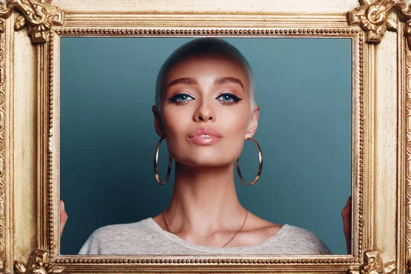 Millenial young woman with short blonde hair holds gilded picture frame in hands behind her face portrait. — Stock Photo, Image