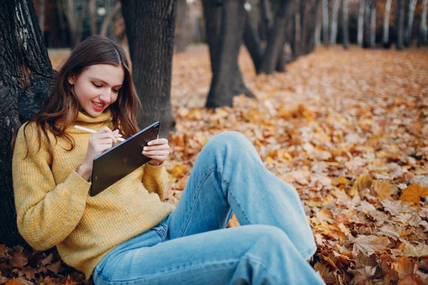 MOSCOW, RUSSIA - PAŹDZIERNIK 19, 2021: Młoda modelka siedząca z tabletem Apple iPad Pro 2021 i malująca ołówkiem w jesiennym parku — Zdjęcie stockowe