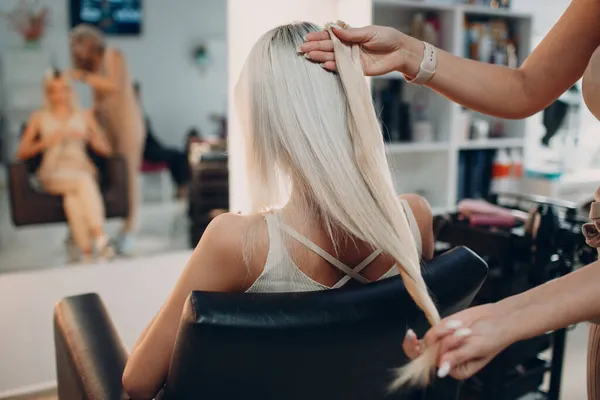 Cabeleireiro feminino fazendo extensões de cabelo para jovem mulher com cabelo loiro no salão de beleza. Extensão profissional do cabelo. — Fotografia de Stock