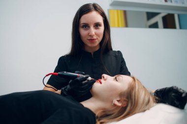 Kozmetik uzmanı genç kadın dudaklarına kalıcı kırmızı makyaj dövmesi yapıyor. Kalıcı dudak dövme prosedürü kavramı.