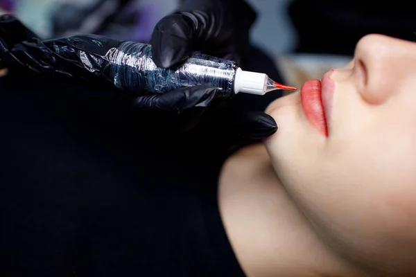 Cosmetoloog het aanbrengen van rode permanente make-up tatoeage op jonge vrouwelijke lippen. Permanente lippen tatoeage procedure concept. — Stockfoto