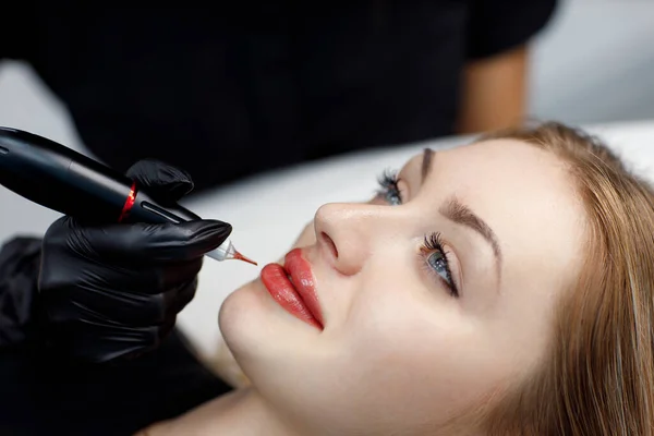 Cosmetologist aplicando tatuagem permanente de pigmento rosa em lábios femininos com máquina de agulha de tatuagem. — Fotografia de Stock