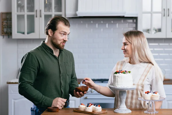 Koki kue manis wanita muda kaukasia dengan suami pacar pria mencoba rasa kue di meja dapur. Kue mangkok dan makanan penutup manis. — Stok Foto