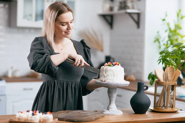 Koki kue manis wanita muda kaukasia dengan kue di meja dapur. — Stok Foto