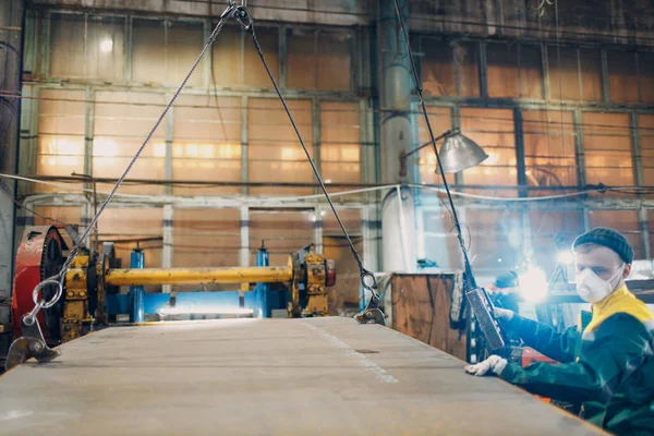 Pekerja mengangkat lembaran logam dengan derek rantai hoist dengan remote controller dan kait di pabrik industri. Stok Foto
