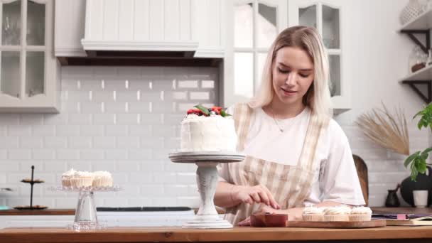 Koki kue manis wanita muda kaukasia dengan kue di meja dapur. — Stok Video