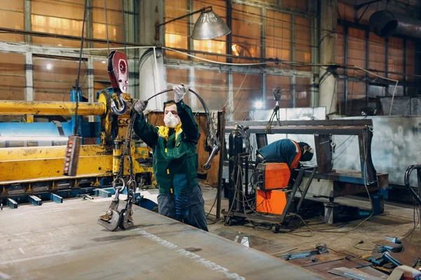 Pekerja mengangkat lembaran logam dengan derek rantai hoist dengan remote controller dan kait di pabrik industri. — Stok Foto