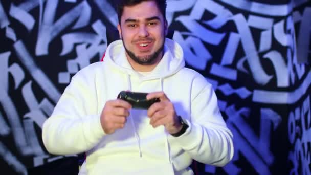 Joven juega en la consola de juegos con gamepad sentado en la silla — Vídeos de Stock