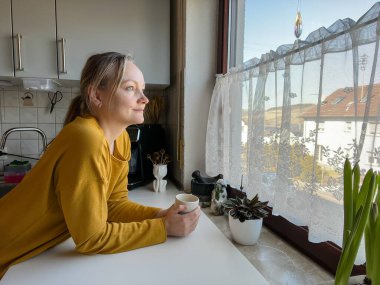 Kadın elinde bardakla pencereden dışarı bakıyor. Mutfakta duruyor.