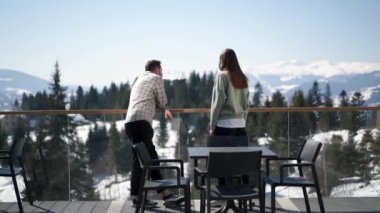 Bir erkek ve bir kız dağlara bakan terasta sohbet ediyorlar.