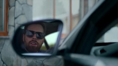 İş adamı karısını arabada bekliyor, dikiz aynasından bakıyor.
