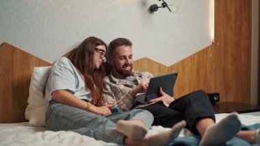 Genç çift otel odasında tabletten video izleyip gülümsüyor.
