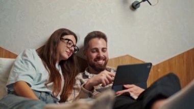 Genç çift otel odasında tabletten video izleyip gülümsüyor.